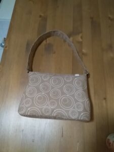 A képen egy világosbarna lapon fehér köröket ábrázoló anyagú felfelé kissé szűkülő trapéz alakú váll/kézi táska, ami nem állítható hosszú.