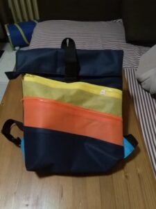 A képen egy roll-top hátizsák látszik, sötétkék alapon ferde cipzáras zsebek vannak rajta narancssárga, fölötte citromsárga színben.