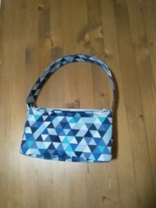 A képen egy apró női kézitáska látható az említett pamutvászon anyagból, ami különböző árnyalatú kék háromszögeket ábrázol.