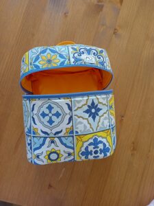 a képen egy sárga-kék marokkói mintás uzsonnás táska látható, ovális alapú henger, tetején apró citromsárga füllel. A táska felső harmadán a három oldalán körbefutó világoskék cizárral csukható. a képen nyitva van, bélése okkersárga egyszínű