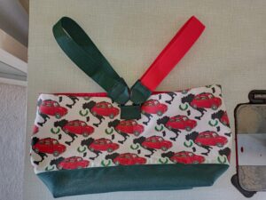a képen egy textil uzsonnás kosár látható üresen, oldalán kiterítve. alja sötétzöld, oldala fehér alapon piros fiat kisautókkal, 2 füle van egyik sötétzöld, másik piros