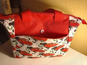 A képen egy női válltáska látszik, alakja felfelé kissé bővülő trapéz. Anyaga fehér alapon piros kis Fiat autók, füle és hevedere piros. elől egy mágneszáras zsebe van, aminek mintája szintén fehér alapon piros autós. fölül cipzárral zárható, pántja állítható hosszúságú pirosheveder. A táskát szemből látjuk.