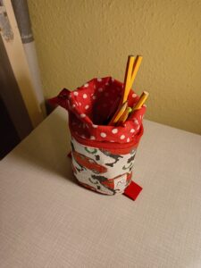 A képen egy tolltartó van, ami fölül cipzáras és a a cipzárat kinyitva a fölső részét rá lehet húzni az alsó felére, ami így elég stabil ahhoz, hogy megálljon az asztalon. A képen éppen nyitva van, ceruzák lógnak ki belőle. kívül piros autók vannak fehér alapon, bélése piros alapon fehér pöttyös.