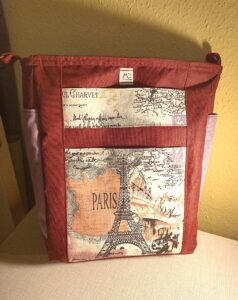 A képen egy nagyalakú álló téglalap alakú válltáskát látunk, ami bordó színű, oldalán mályva színű nyitott zsebek palack tárolására. Az elején egy nyitott és egy mágneszáras zseb van egymás fölött, ami Párizs ihletésű mintás anyag Eiffel toronnyal, Diadalív, francia feliratok. a táska állítható pántja bordó színű. A táska szivaccsal bélelt, laptop szállítására tervezve. A táskát szemből látjuk.