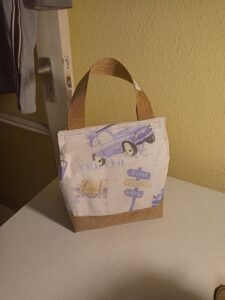 a képen egy uzsonnás táska látható szemből. anyaga fehér alapon halvány lila fiat autóval és olaszországi útjelző táblákkal. a táska alja és füle világosbarna. a tetején belül tépőzárral csukható