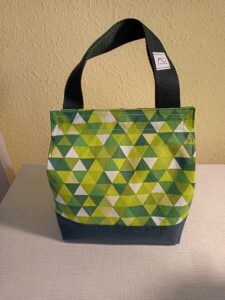 a képen egy uzsonnás táska látható szemből. anyaga hefér és különböző árnyalatú zöld háromszögekkel mintás. a táska alja és füle sötétzöld. a tetején belül tépőzárral csukható