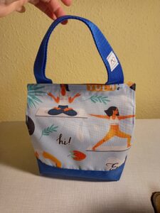 a képen egy uzsonnás táska látható szemből, a tetején tépőzárral csukódik. anyaga világoskék alapon női alak jógapózokat mutat be. a táska alja és füle királykék