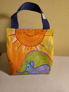 a képen egy uzsonnás táska látható szemből, a tetején tépőzárral csukódik. anyaga gyerekrajz mintás, egy hatalmas napot és egy lila elefánt tetejét látjuk