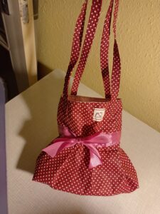 A képen egy hosszú vékonypántos, bordó alapon apró pöttyös mintájú pamutból készült kistáska van, ami elölről úgy éz ki, mintha egy kis ruha lenne, a táska fele fölül egyenesfalú, közepén (derekán) rózsaszín masnival, az alsó fele bővülő, mintha egy pörgős szoknya lenne.