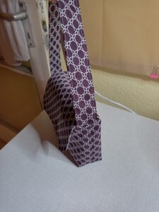 A képen egy apró női kézitáska van. Anyaga bordó alapon fehér egymásba fonódó lekerekített négyzetek, teteje lehajlik és patenttel záródik. A táska tényleg apró, maximum egy mobil, egy átlagos méretű pénztárca és egy csomag zsebkendő fér bele. A táskát oldalról látjuk.