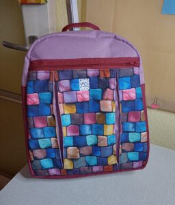 A képen egy lekerekített tetejű hátizsákot látunk, aminek alja és oldala bordó, teteje és eleje fölül mályva színű, elejének nagyját egy cipzáras zseb takarja ami tarka kockakő mintás. A táska fölül cipzárral záródik, amely takarásban van, így viselés közben sem látszik.