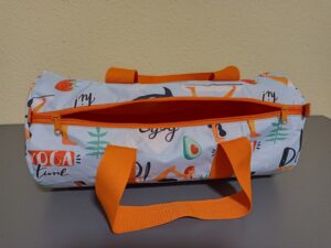 A képen egy henger alakú sporttáska van, alja, pántja és cipzára narancssárga, a táska teste világoskék alapon jóga pózokat bemutató nőik alakok. az alakbőrtónusa szintén narancsszínű, ezzel harmonizál a többi narancssárga rész. a táskát a vége felől látjuk, nyitva van, bélése is narancssárga