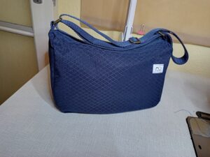 A képen egy apró női válltáska látható, állítható hosszúságú vékony pánttal. A táska sötétkék alapon anyagában rombuszmintás. A táskát szemből látjuk. Felül cipzárral zárható.