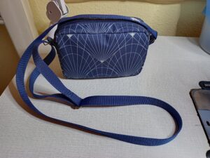 A képen egy apró női válltáska látható, állítható hosszúságú vékony pánttal. A táska sötétkék alapon boltívek és sugár irányú vonalakkal mintázott. A táskát szemből látjuk. Felül cipzárral zárható.