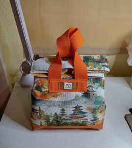 A képen egy japán ihletésű uzsonnástáskát látunk. Külső anyagán különböző japán templomokat és erdőt, hegyet látunk rajzolva, a táska alja és fülei narancssárgák (mivel az épületek fa részei narancssárga színűek). A táska fülei is egyedi megoldással, japán stílusban úgy van megoldva: egyik füle kisebb, ezen van átbújtatva a nagyobb méretű fül, amit csuklóra téve szoktak vinni. A tásja tetejét le kell hajtani ahhoz, hogy a füleket át tudjuk húzni egymáson. ezen a képen csukva, szemből látjuk a táskát.