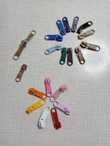 a képen cipzár húzókákat (kocsikat) látunk fehér, citromsárga, narancssárga, rózsaszín, fáradt rózsaszín, halványlila, bordó, sötétzöld, világoskék, királykék, élénk kék, sötétkék, szürke, fekete, világosbarna, középbarna, sötétbarna és fém színekben