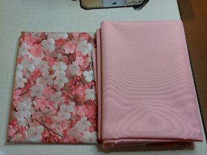 a képen gyöngyvászon anyagok vannak egymás mellett, egyik fehér, rózsaszín cseresznyevirágokkal díszítve, a másik pedig fáradt-rózsaszín, színben passzol a mintáshoz