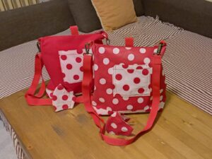 A képen 2 női táska van, amik válltáskaként és hátizsákként is használhatók. egyik piros egyszínű, elől zsebe fehér alapon piros nagy pöttyös, előtte egy ugyanilyen maszk, a másik táska piros alapon fehér nagy pöttyös, a táska előtt egy ilyen mintás maszk