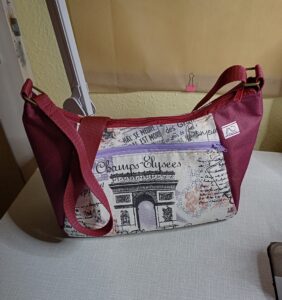 A képen egy felfelé bővülő női válltáskát látunk, ami bordó színű, és az elején cipzáras zseb van, ami Párizs ihletésű mintás anyag Diadalívvel, hátoldalán pedig Eiffel toronnyal. A táska pántja állítható, bordó színű