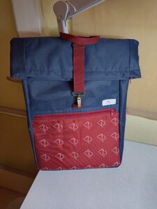 A képen egy sötétkék rolltop hátizsák látható, elején egy bordó cipzáras zsebbel, amin fehér színnel az M. Dina Bags cicás logója van 4 sorban, 45 fokban ferdén.