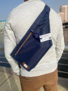 A képen egy sötétkék cross-body táska látható, aminek formája egy függőlegesen félbevágott gitárra hasonlít. A cipzárak barna színűek. A táska éppen keresztbe vetve egy férfi hátán látható.