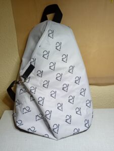 A képen egy fehér alapon fekete mintás hátizsák látható, az elején egy ferde cipzárral. A minta a cicás MD logó ferdén az anyagon. A táska formája kissé szokatlan, az alja szélesebb, a teteje szűkül, lekerekített. Keskeny fekete pánton karabínerrel lehet rögzíteni a cipzár alján lévő D karikánál. Így lehet rögzíteni a táska testét, ha nincsen tele.